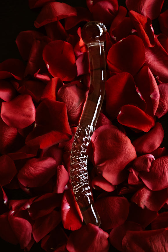 A clear borosilicate glass wand lying on a bed of red rose petals. The glass wand has a bulbous end and textured ridges along its length.