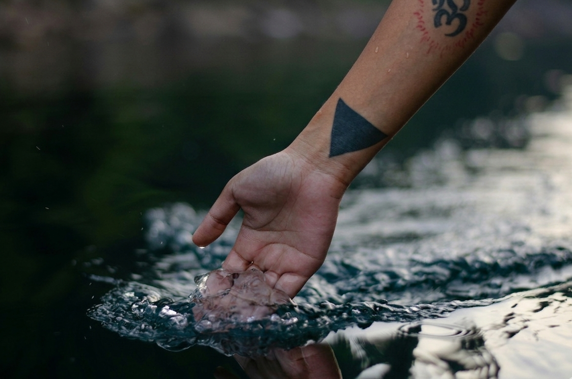 Hand with a triangle tattoo touching water, with a blurred natural background